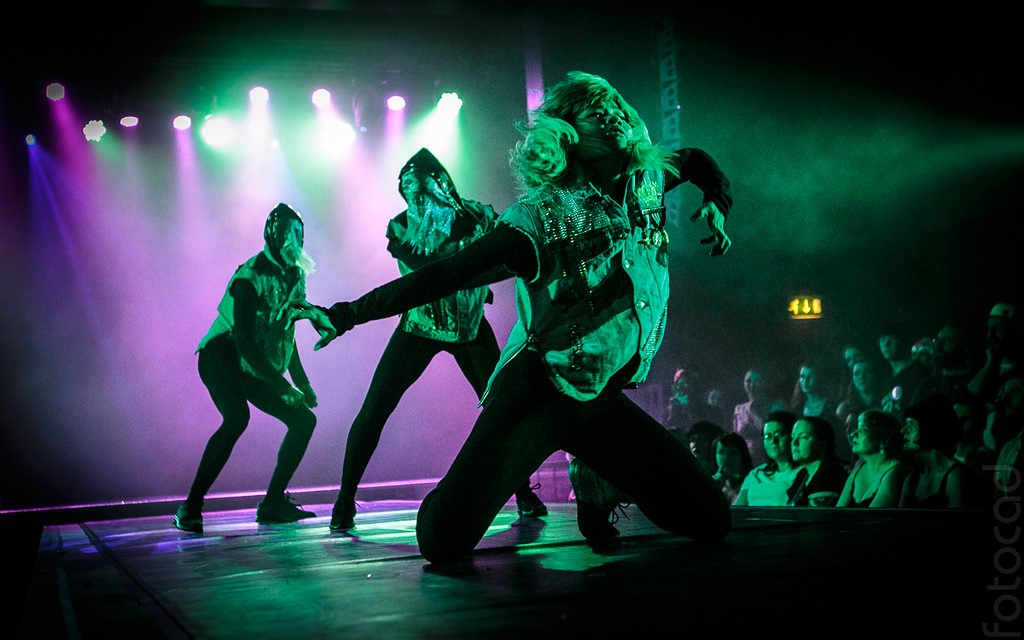 Three dancers onstage under purple and green lighting. The foremost is on their knees with their arms spread out dramatically.